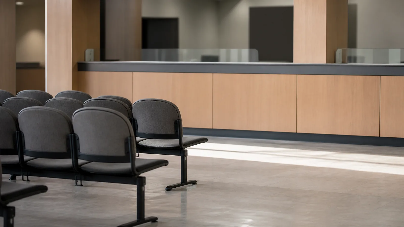 The interior of a quiet public service office with empty chairs in rows and a plain counter, winter light falling in bands across a pale floor.