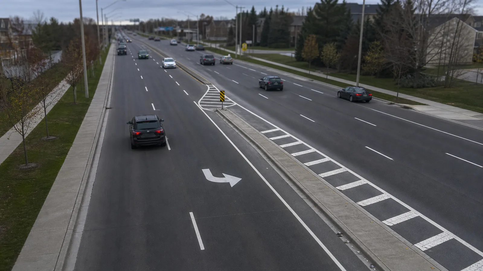 A multi lane road with a marked left turn bay and a painted island, seen from a footbridge above moderate traffic under flat daylight.