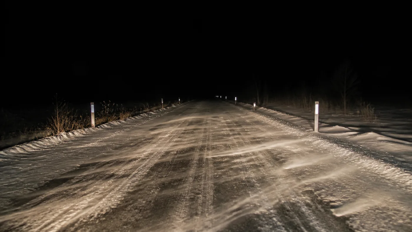 A rural road at night lit only by headlights, snow drifting across the surface and the verge markers catching the light.