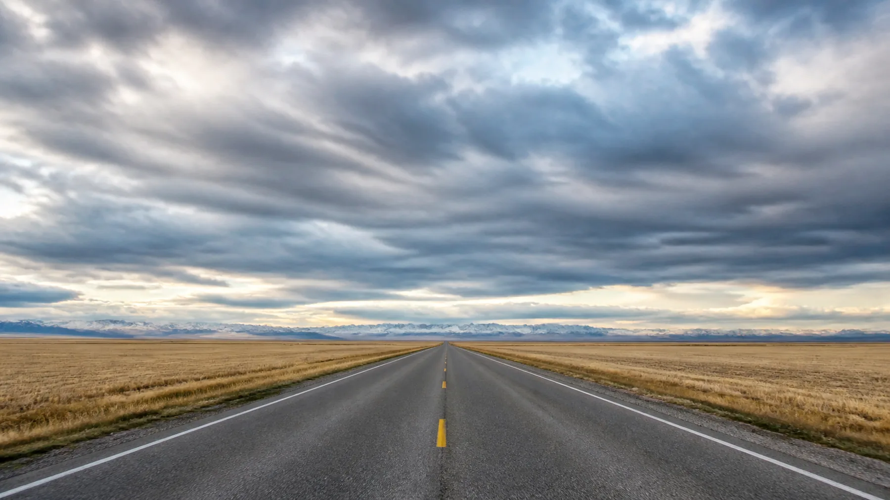 A straight prairie highway crossing stubble fields under an immense bank of cloud, with a mountain range as a pale band on the far horizon.