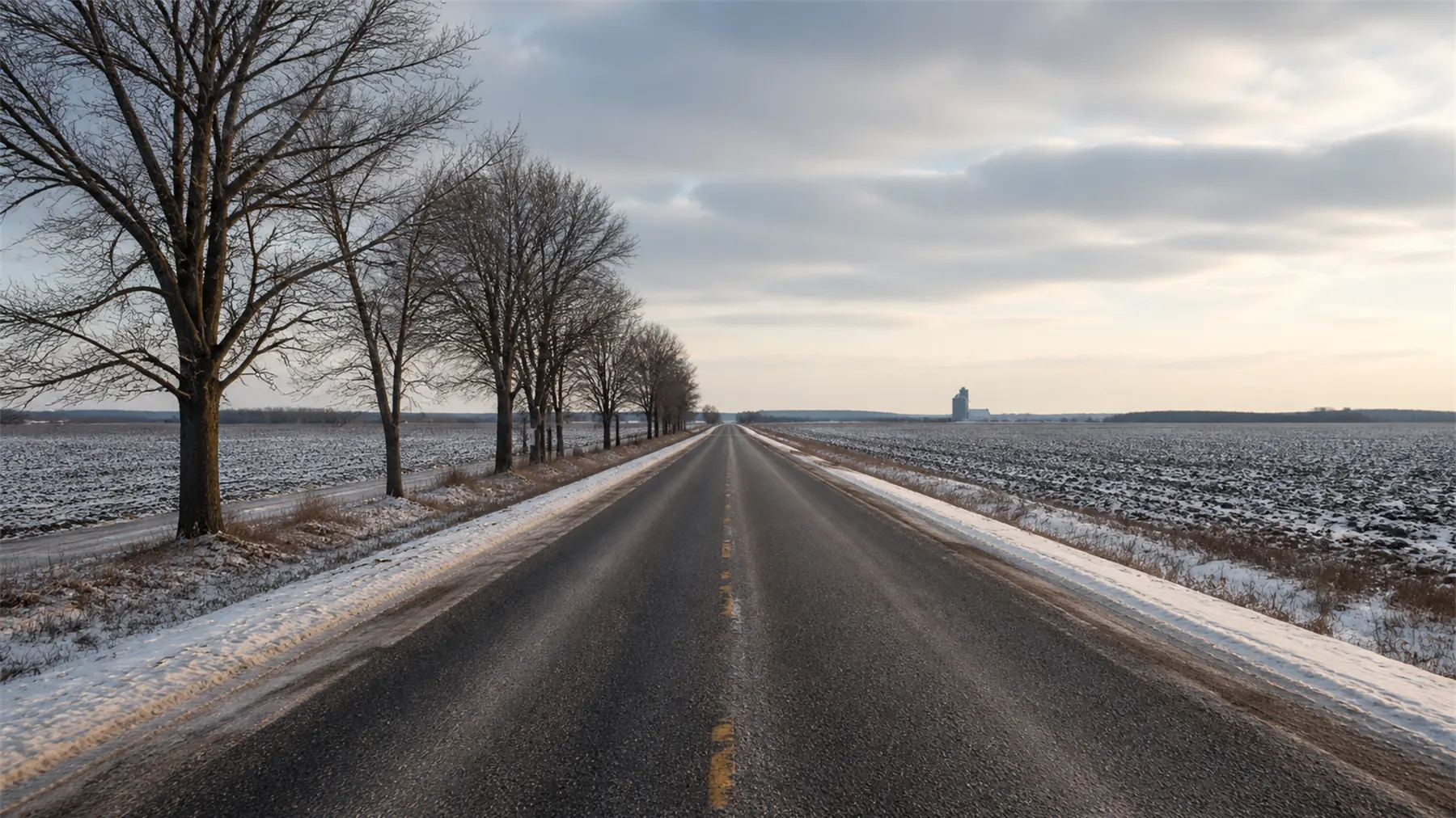 A straight rural road running to a low horizon between snow dusted fields, bare maples along one side and a grain elevator silhouetted far off.
