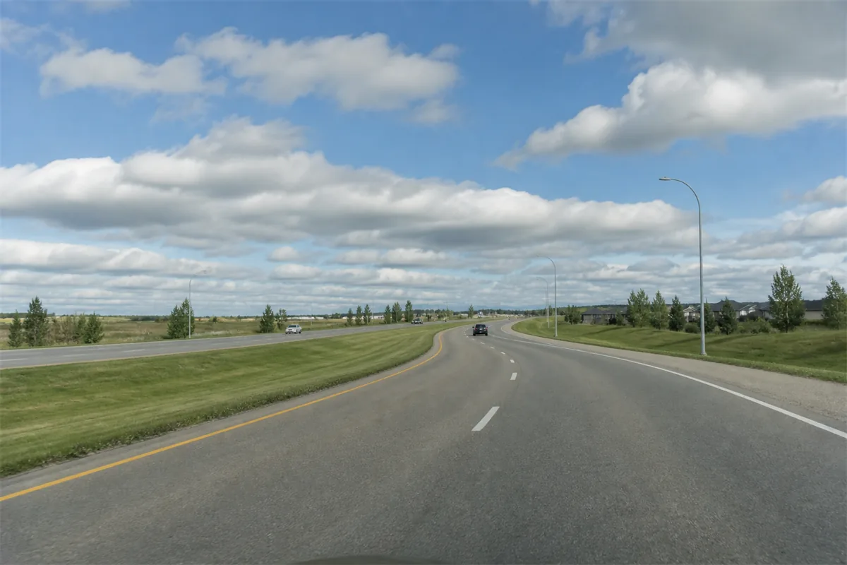 A driver view of a wide arterial road with a shallow bend and a broad grass median, big sky and scattered cloud overhead.