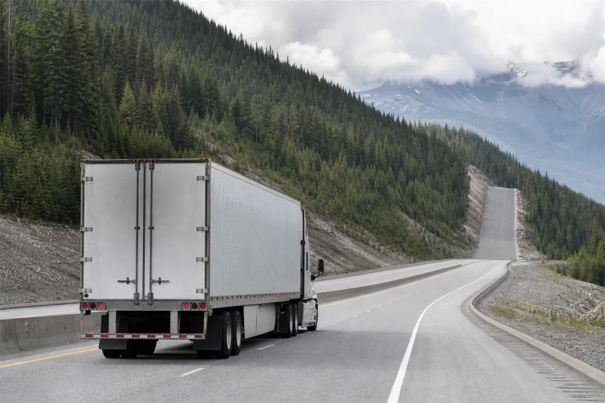 A tractor trailer climbing a long mountain grade on a divided highway, forested slope rising to the left and the gravel of a runaway lane visible ahead.