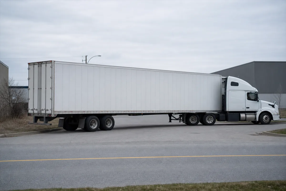 A tractor trailer taking a wide right turn from a two lane road into an industrial entrance, seen from the opposite curb under overcast light.